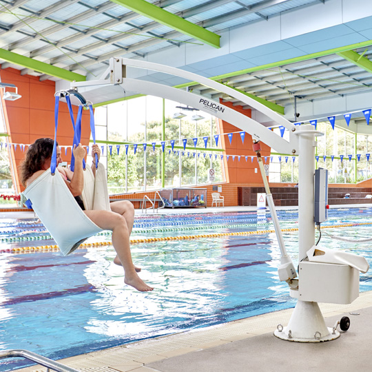 Woman sitting in pelican pool hoist being assisted into an indoor pool